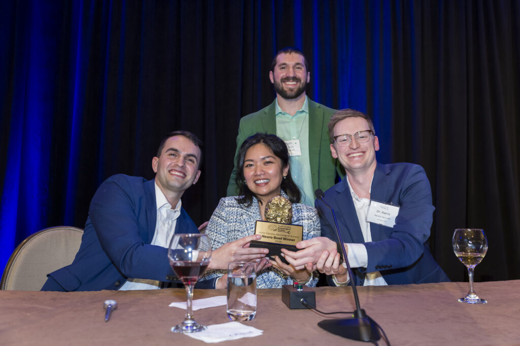 Two seated men and one woman hold a trophy featuring a brain, with one man in the background